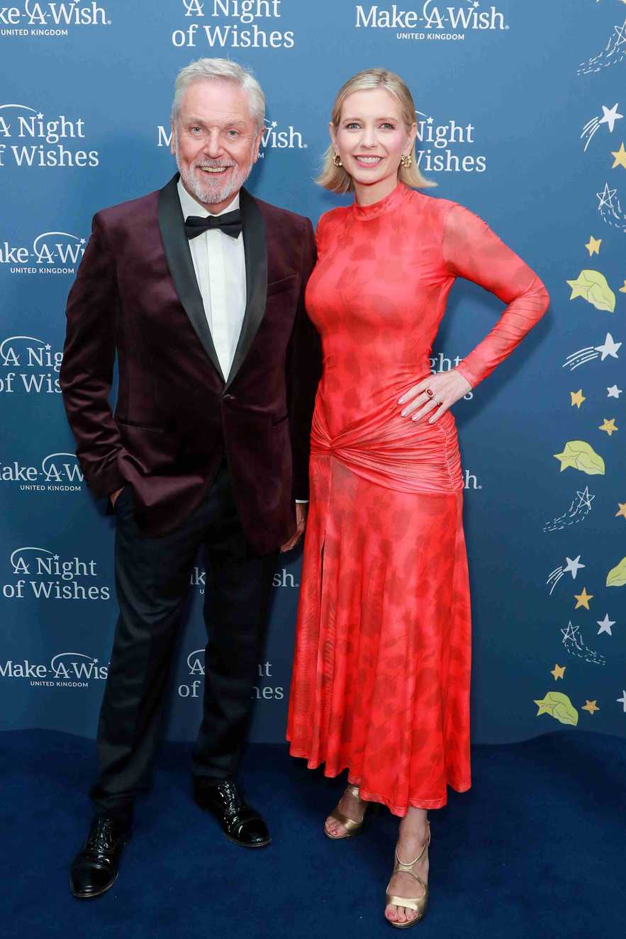 A man dressed in a maroon velvet tuxedo and a woman dressed in a red dress pose on a blue carpet.  Behind them is a backdrop showing the Make-A-Wish brand