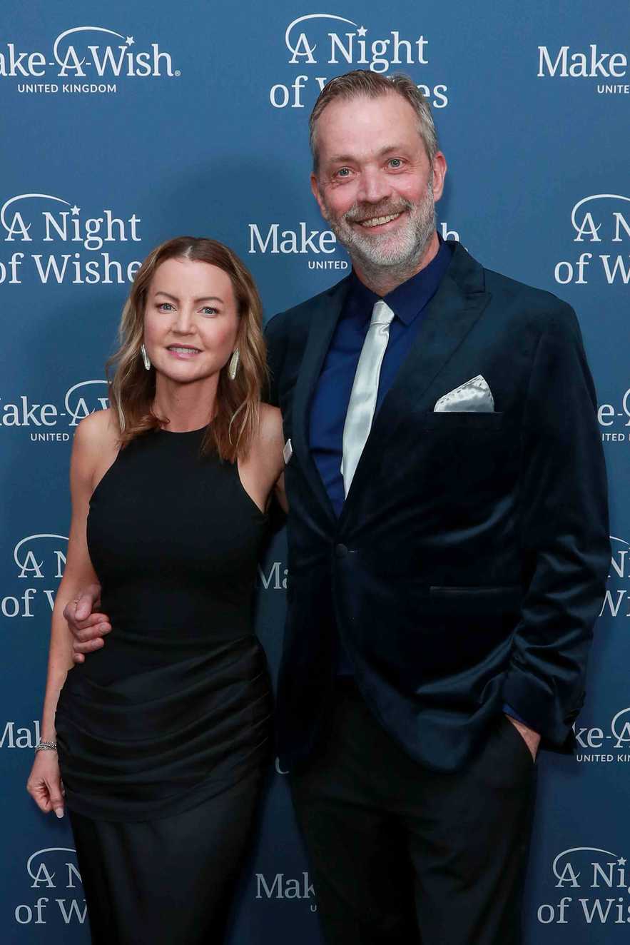 A man and a woman are standing smiling at the camera. The woman is wearing a bacl dress and the man a blue suit with a silver tie.