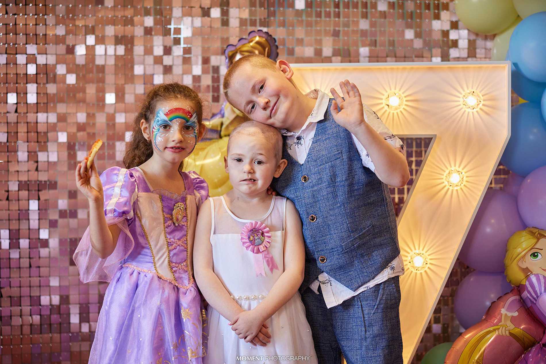 Ryleigh and two friends wave at the camera as they stand in front of a giant illuminated number seven.