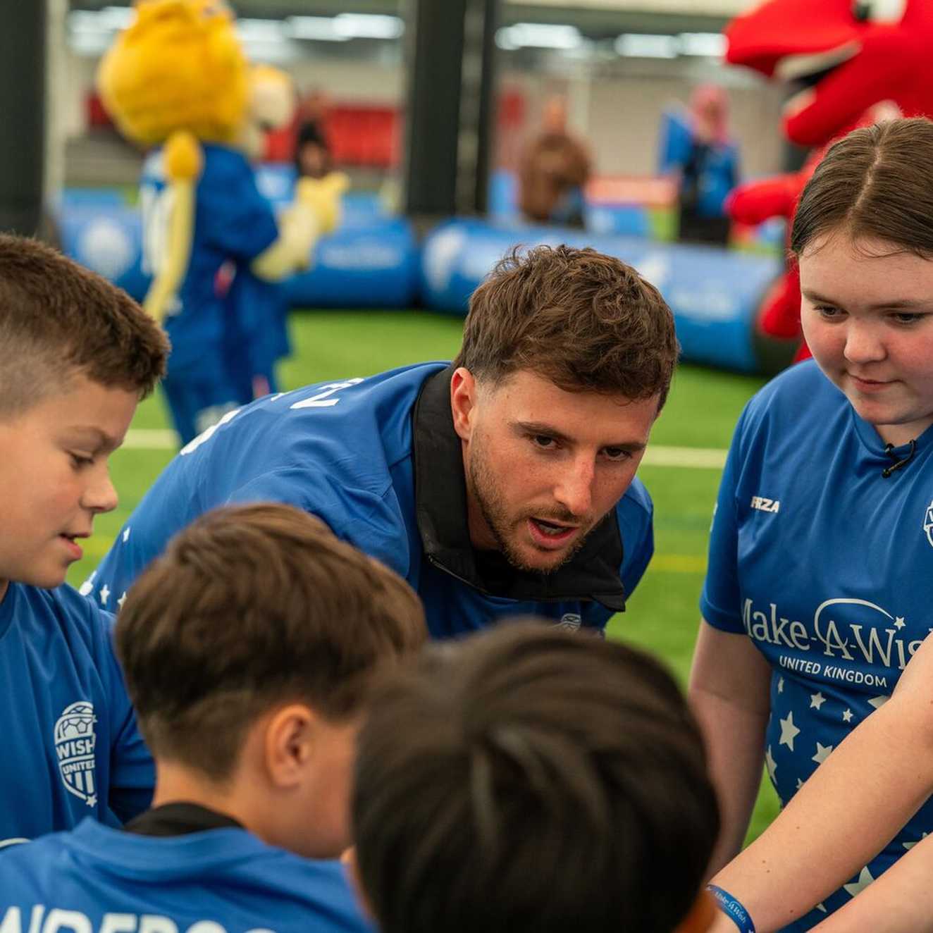 Mason Mount gives a team talk to the children. They are all putting their hands together.  Everyone is wearing a Make-A-Wish, Wishes United football top.  Behind them is a football pitch.