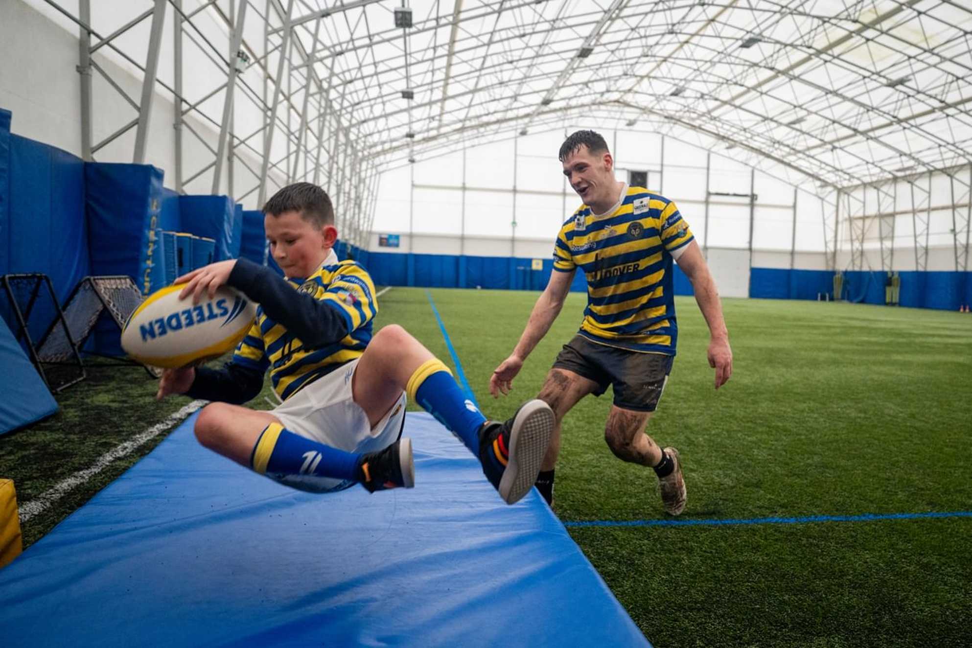 Callum dives on to a crash mat as if scoring a try, with Josh Thewlis behind him. Both are in full Warrington Wolves kit.