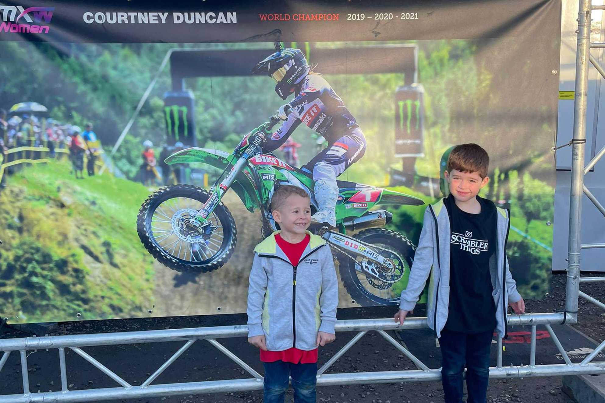 George and his brother, Charlie standing in front a large sign featuring Motocross rider, Courtney Duncan.