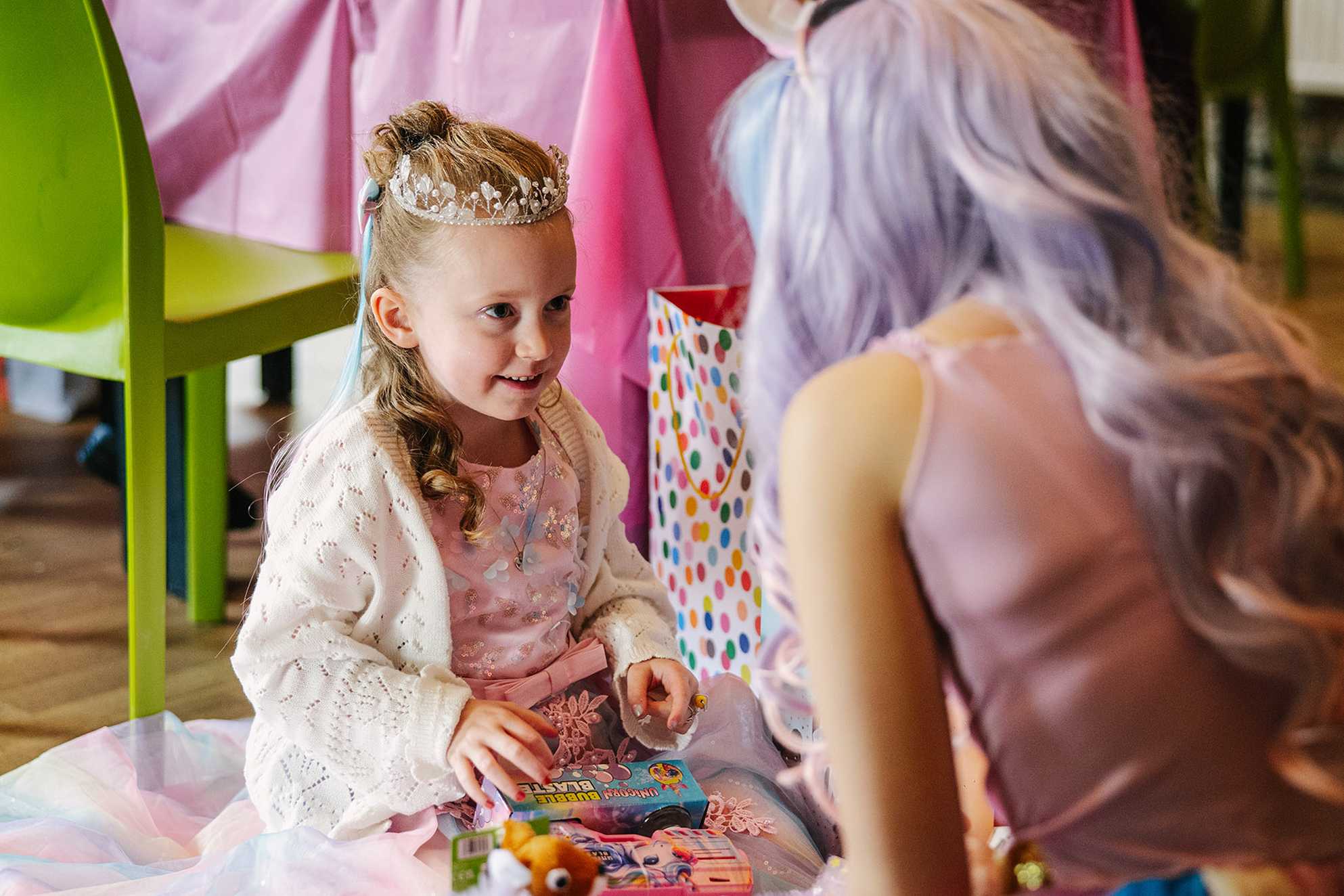 Penny sits down having opened some toy presents, looking and smiling at a grown up princess character