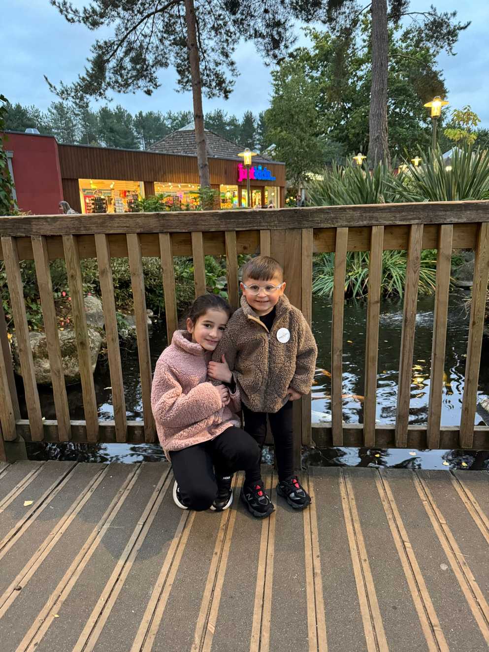 Dzemil is standing smiling next to his big sister who is knelt beside him. They are standing on a bridge with water behind them. Dzemil is wearing a white and blue Make-A-Wish UK badge.