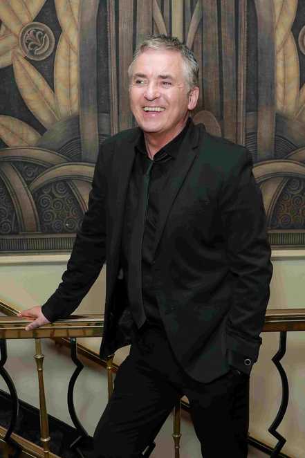 A man is stood smiling in front of a banister.  He is wearing a black suit.