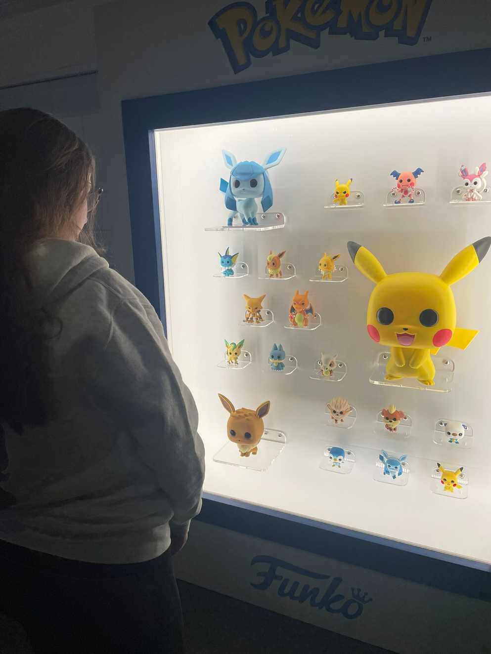 Amelia is stood in front of a brightly lit display cabinet with a collection of Pokémon Funko Pop figures displayed. In the centre, there's a large Pikachu.