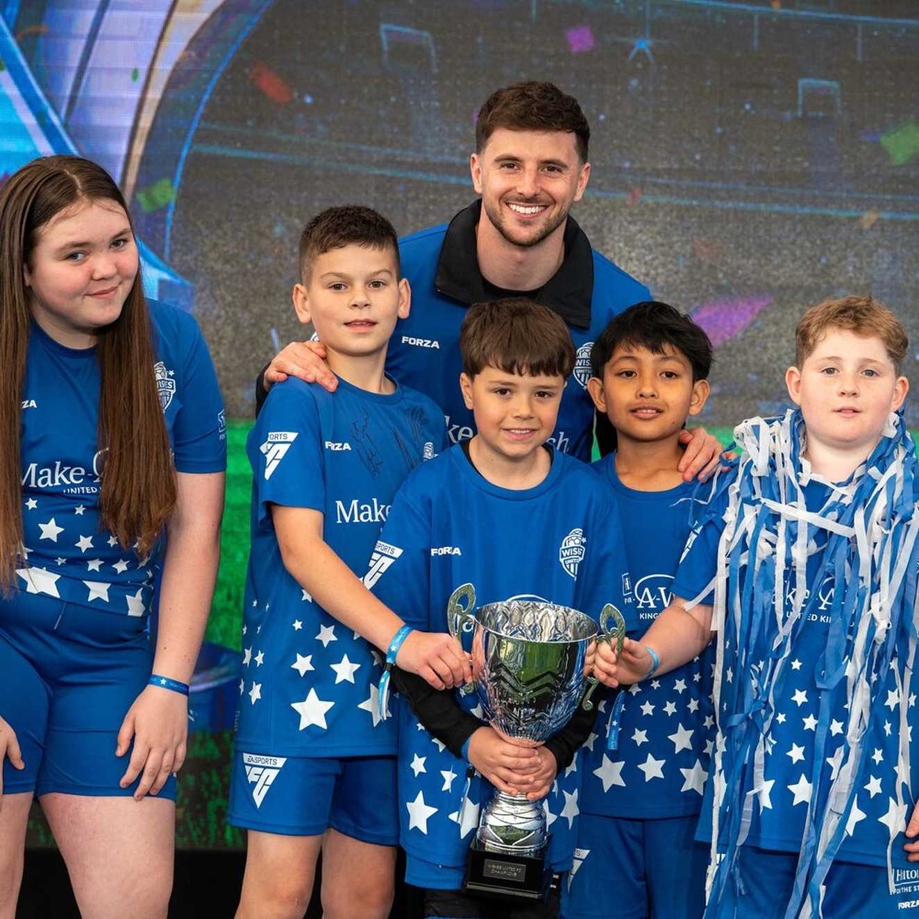 The footballer Mason Mount is smiling and celebrating with five young people.  They are holding a trophy and one boy is covered in ticker tape.  They are all wearing the blue Make-A-Wish football strip.