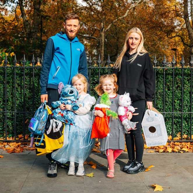 Demi stands on a path next to her mum, twin sister and dad, whilst they carry lots of shopping. Demi and her sister are holding toys including a green The Grinch toy, a pink cat and a Blue Stitch toy.