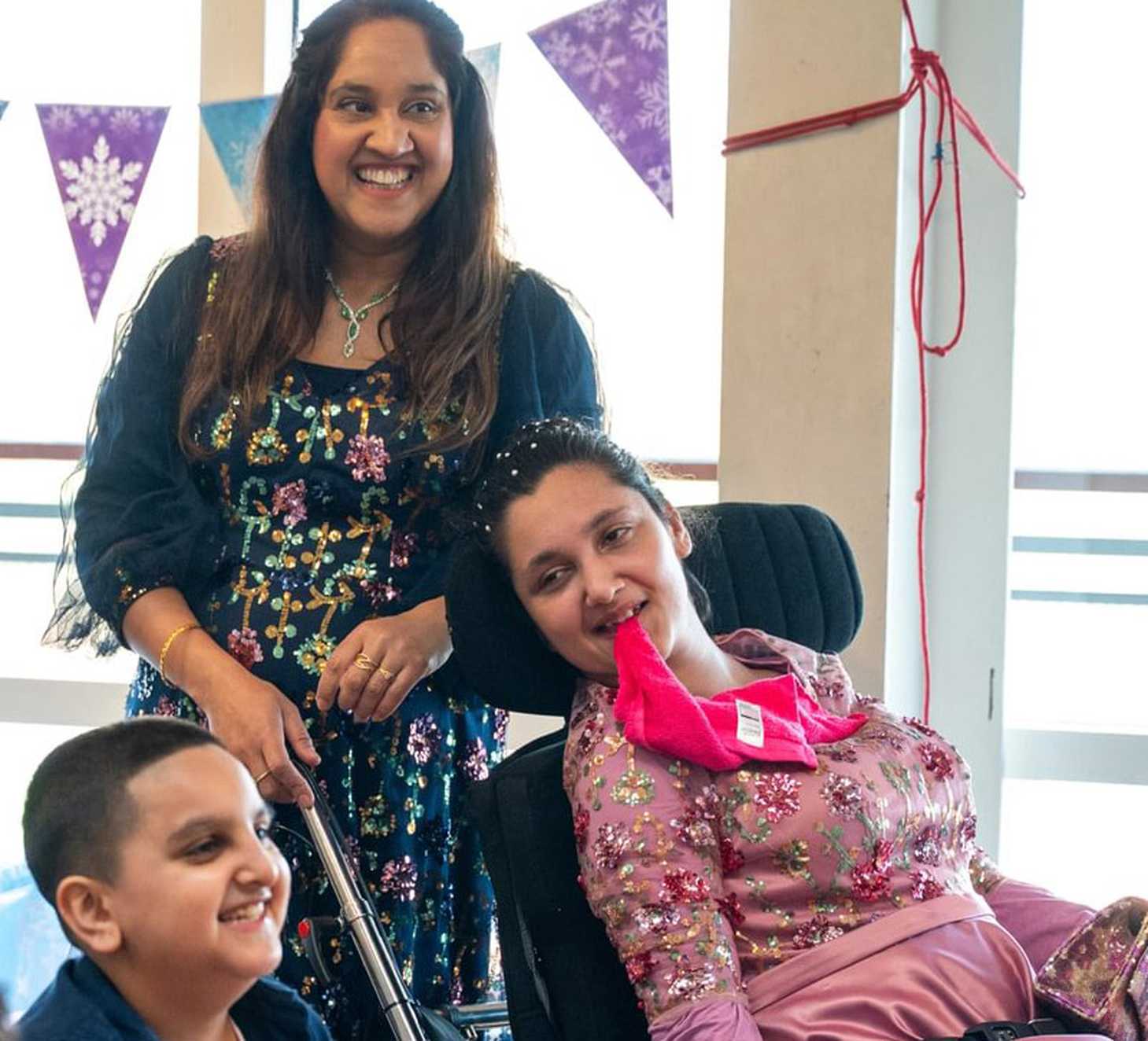 Sumaiya and her family are smiling at the party. Sumaiya is sat in her wheelchair wearing a pink dress and chewing a pink scarf.