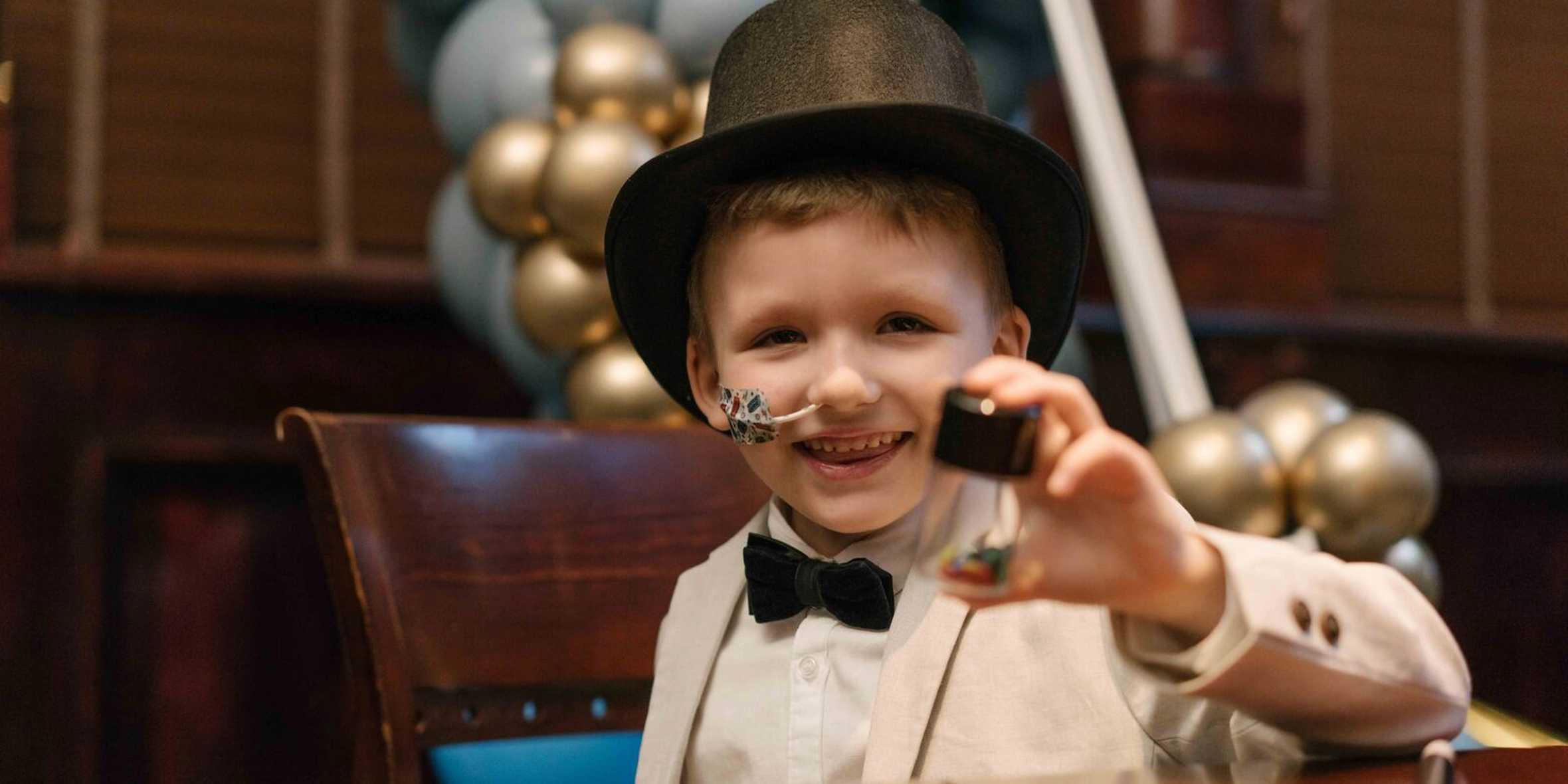 Charlie is smiling at the camera dressed in a magicians outfit, holding a magic device.