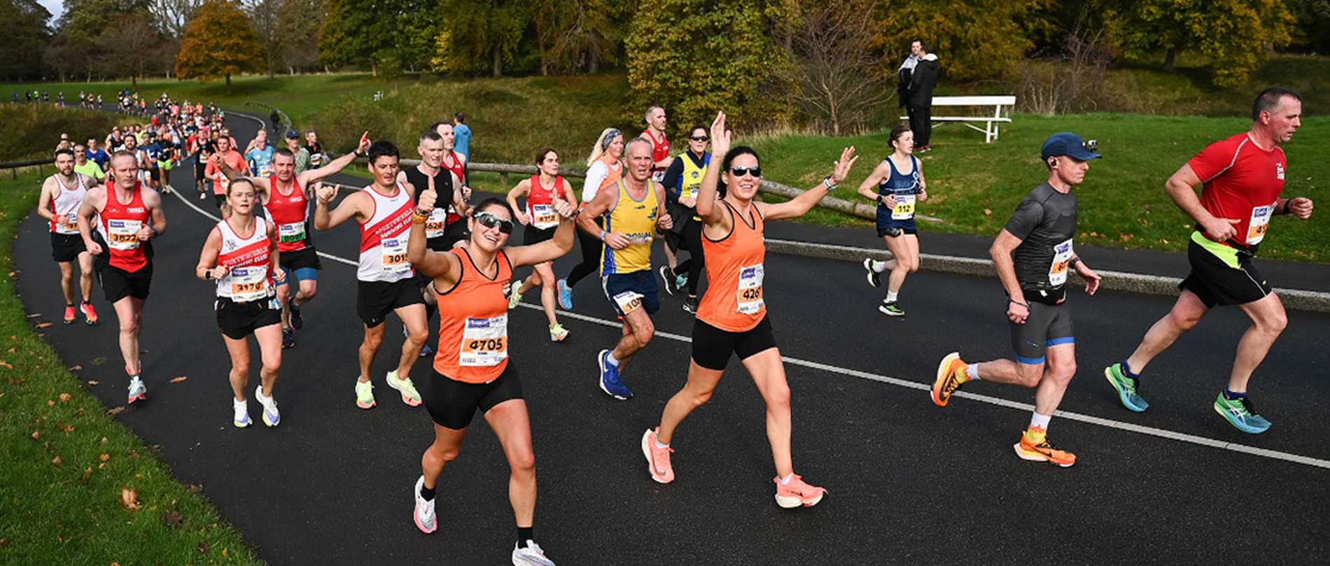 A group of Dublin Marathon runners making their way through one of the city's parks, with two participants in orange vests waving at the camera as they pass.