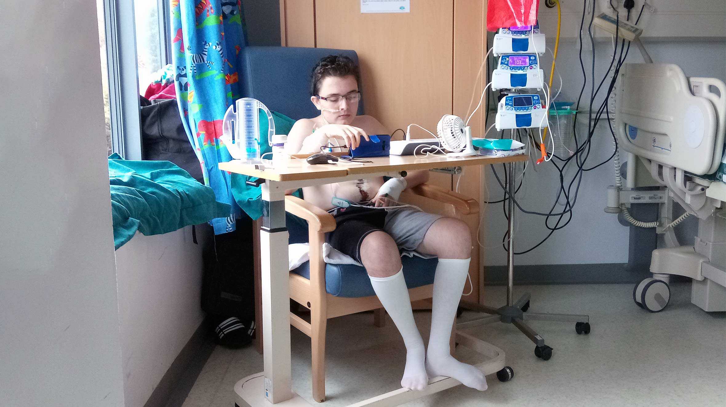 Evan sitting in a chair in a hospital ward, attached to monitoring equipment after his operation.