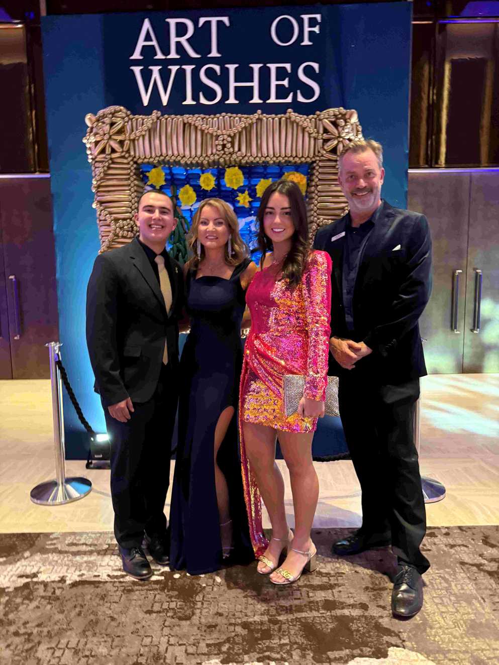 A group of four people dressed formally stand together at the "Art of Wishes" event. They are posing in front of a decorative backdrop adorned with blue and gold accents.