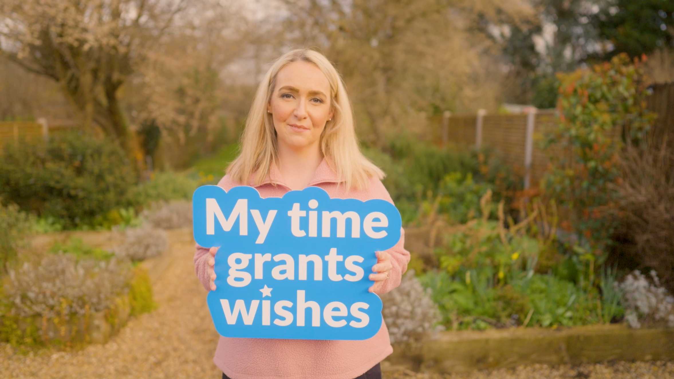 A screenshot of a female Make-A-Wish volunteer in a garden holding a sign saying 'My time grants wishes'