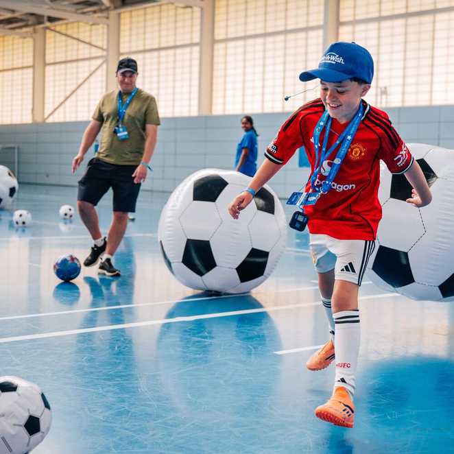 A boy is smiling as he goes to kick a football.  He is wearing a Make-A-Wish cap and is surrounded by other large footballs.