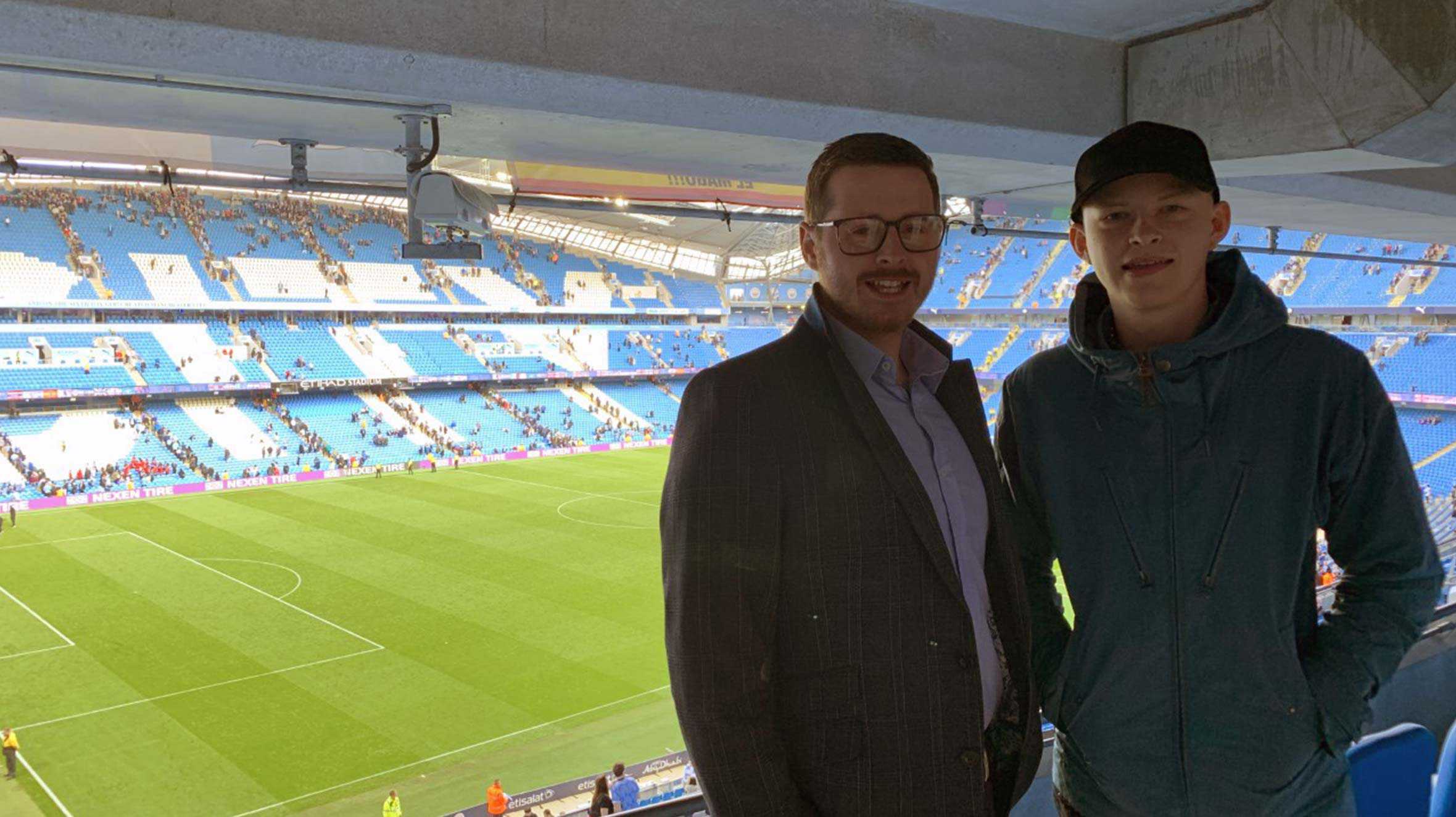 Harrison and Josh in their VIP box facing the camera and smiling, with the football pitch behind them.