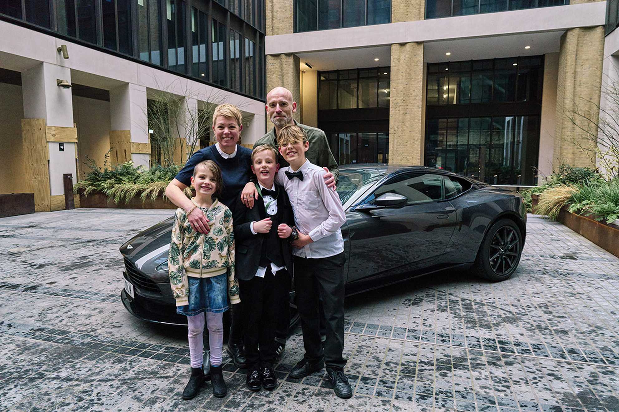 Rudy stands with his mum, dad, sister, and brother in front of a dark grey Aston Martin. Everyone is smiling, dressed smartly, with a modern building featuring tall windows behind them.