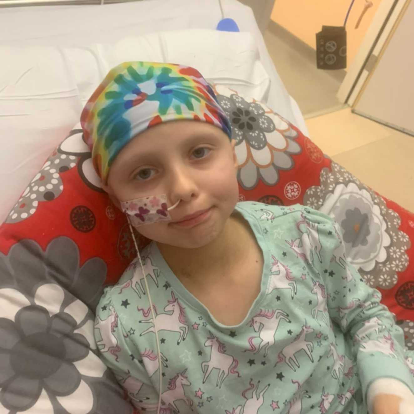 Rosie is sat in a hospital bed with a red, grey and white pillow. She is wearing unicorn themed pyjamas, with a colourful bandana and a feeding tube with butterfly tape secured on her face.