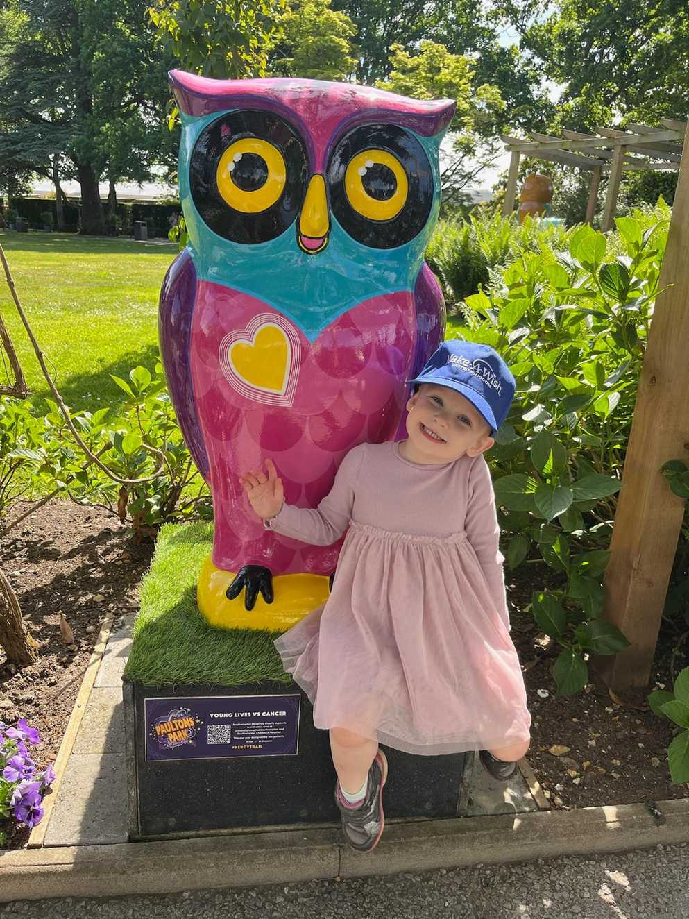 Annalena sits on a box in front of a colourful bird statue. On the birds chest is a yellow heart. Annalena is smiling with a pink dress and a blue Make-A-Wish UK hat on. In the background there are trees and bushes.