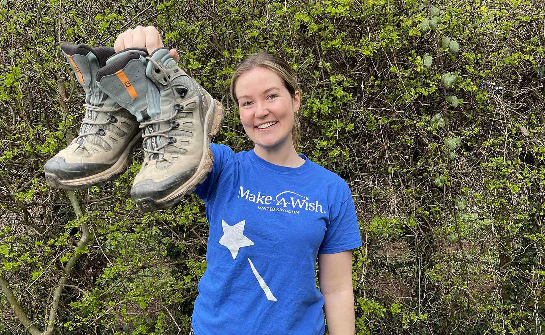 Team member, Callie wearing a blue Make-A-Wish t-shirt and holding up a pair of hiking boots.