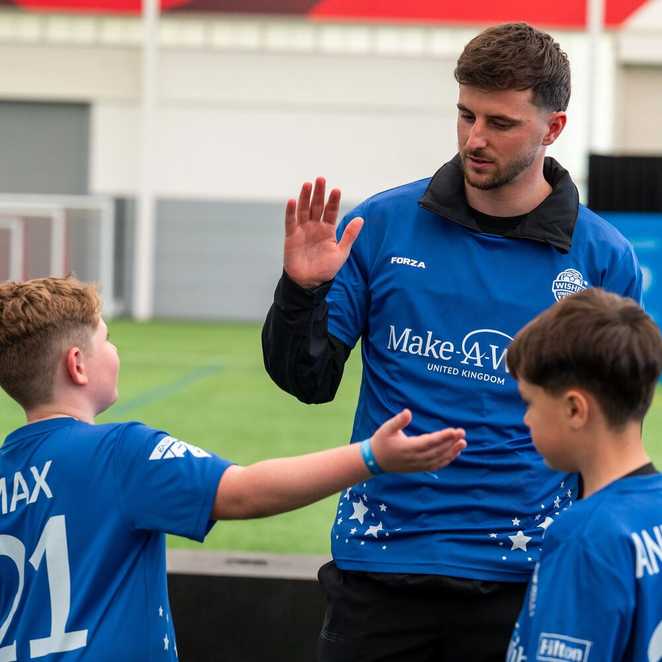 The footballer Mason Mount is high fiving a boy called Max. Behind them is a football pitch and another young boy is standing next to them.  they are all wearing Wishes United football strips.