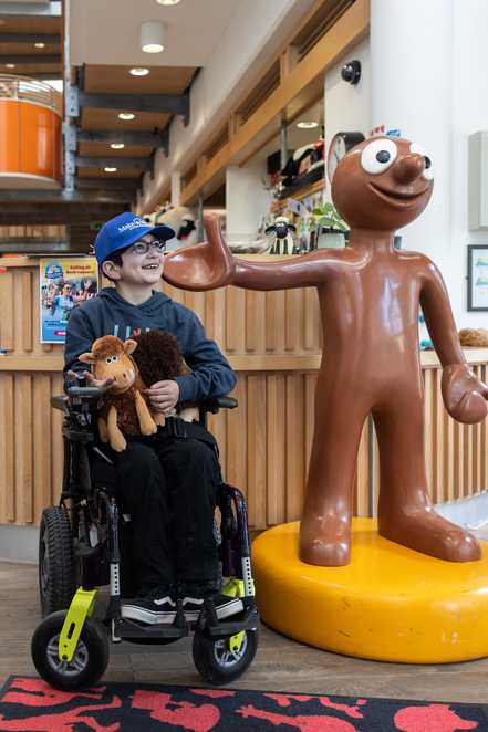 Mert is smiling, looking to his left in his wheelchair. He is wearing a blue Make-A-Wish UK cap and on his lap are two Early Man toy characters, Dug and Hognob. He is in the reception area of Aardman studio, beside a life-sized model of Morph.