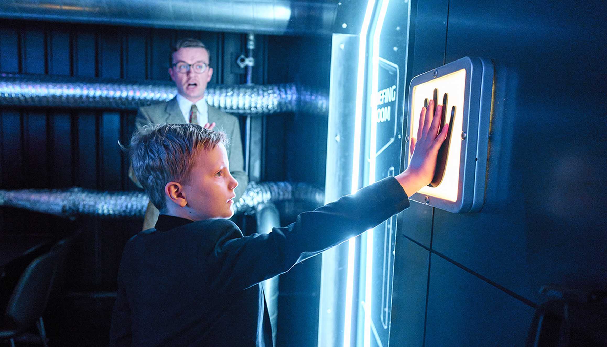 Rudy holds his hand up to a glowing blue hand scanner mounted on a door. He’s dressed in a black suit. Behind him, a man in a beige suit and glasses watches the scan. The background features a dark wall with metallic silver vents.