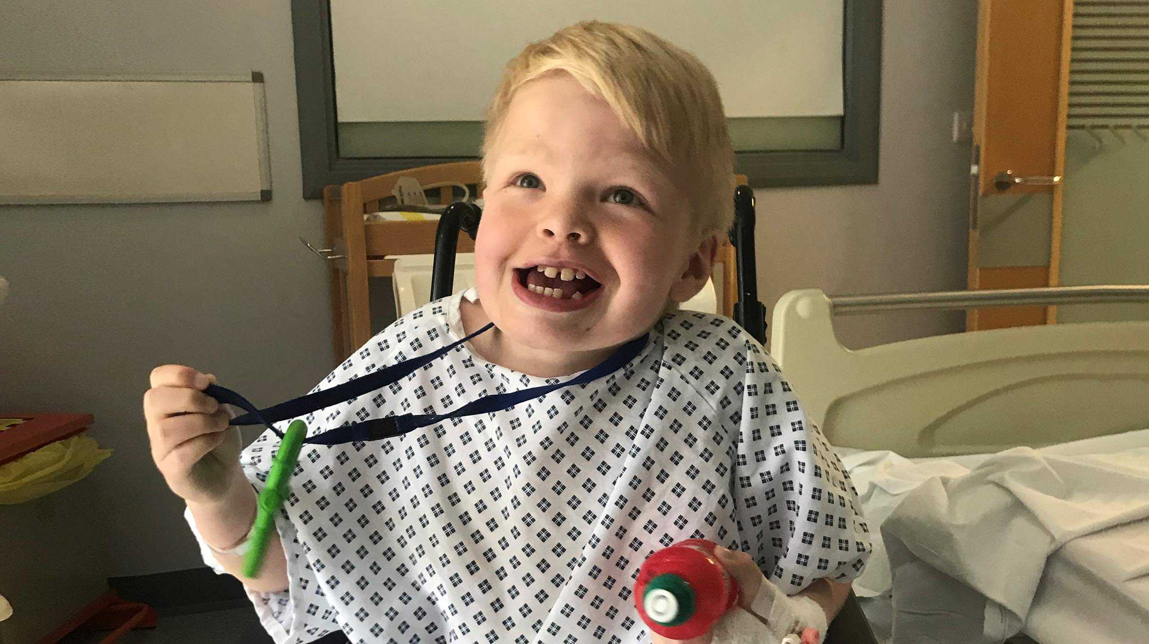 Jack in hospital, wearing a hospital gown with a plastic dummy around his neck and a cheeky smile on his face.