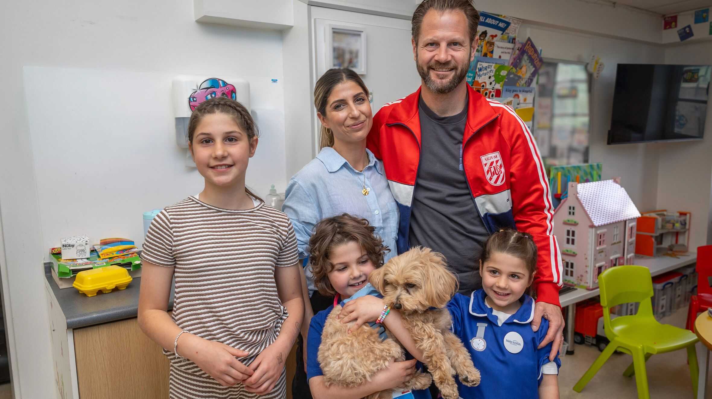 A family (Arabella's) are posing for a picture. Top row from the left is an older girl wearing a stripey dress, then Arabella's mum and her father.  Front row is Arabella holding the family dog and then her younger sister wearing a nurses uniform.