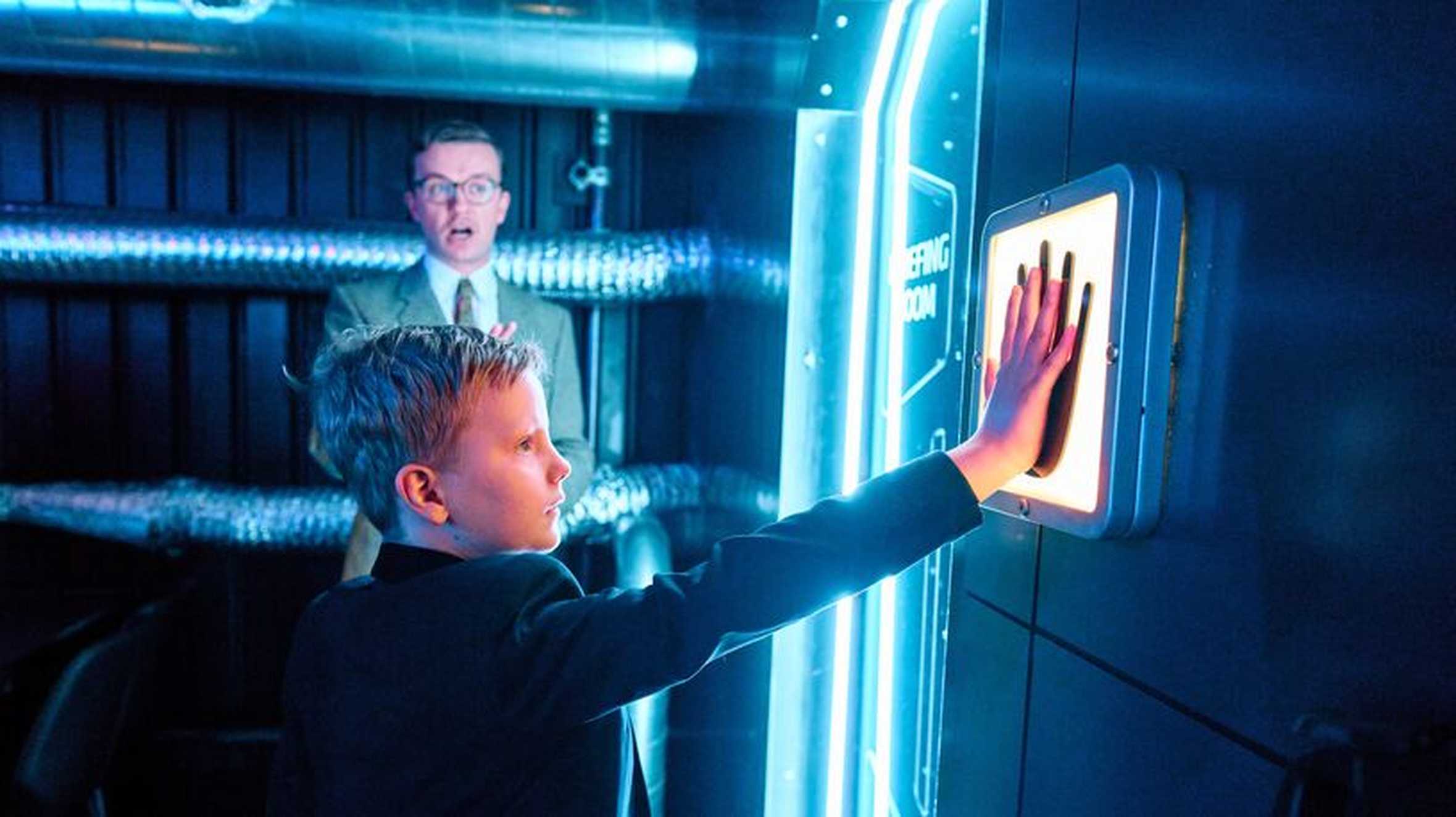 Rudy holds his hand up to a glowing blue hand scanner mounted on a door. He’s dressed in a black suit. Behind him, a man in a beige suit and glasses watches the scan. The background features a dark wall with metallic silver vents.