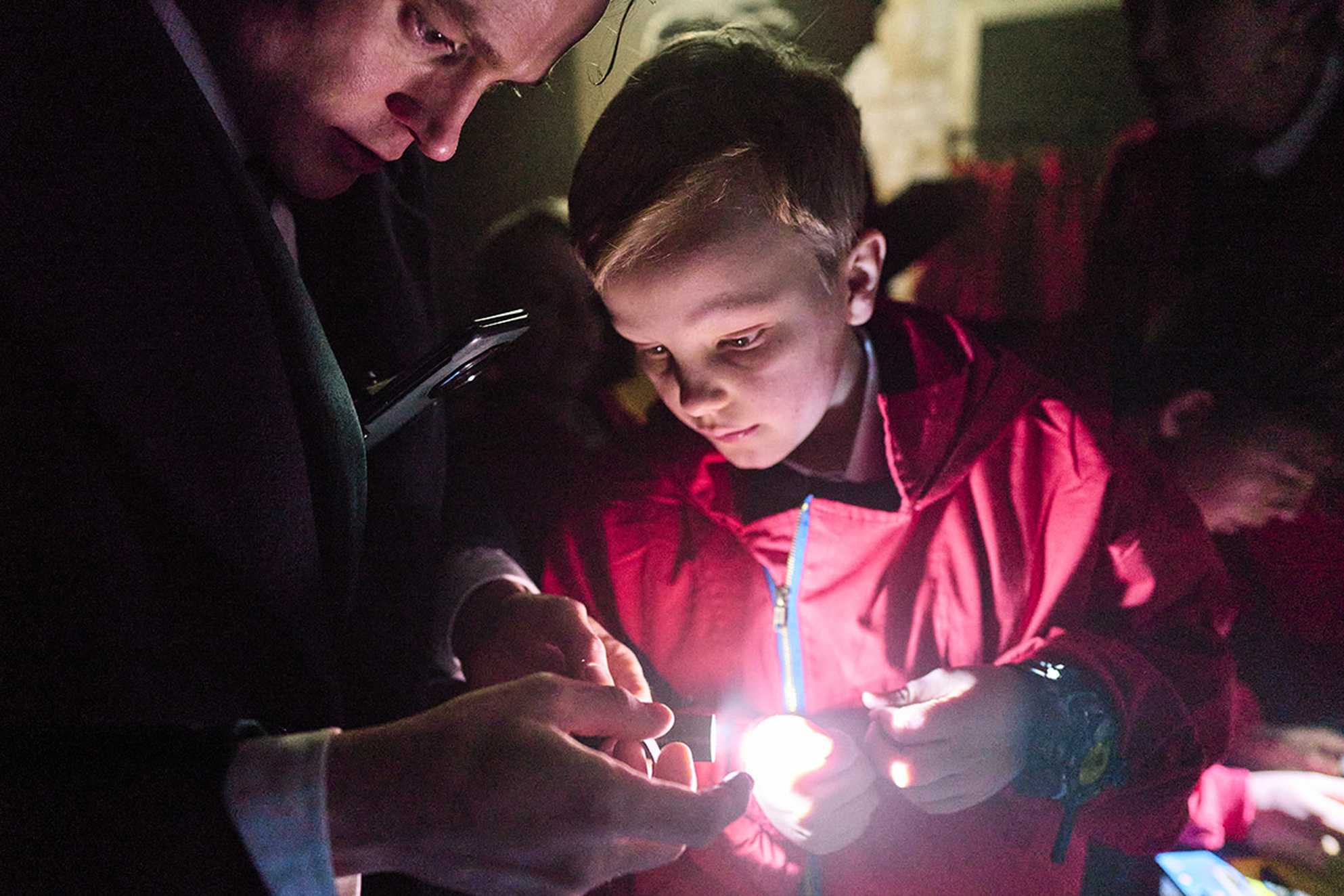 Rudy’s face glows in the light of a flashlight held by a suited man. He’s wearing red overalls and a spy watch, focused intently on the light.