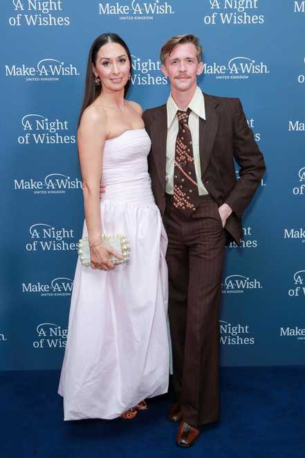 A man and a woman are standing on a blue carpet.  The woman on the left is wearing a pink dress and the man on the right is wearing a brown suit.  Behind them is a wall with the Make-A-Wish A Night of Wishes logos on it.