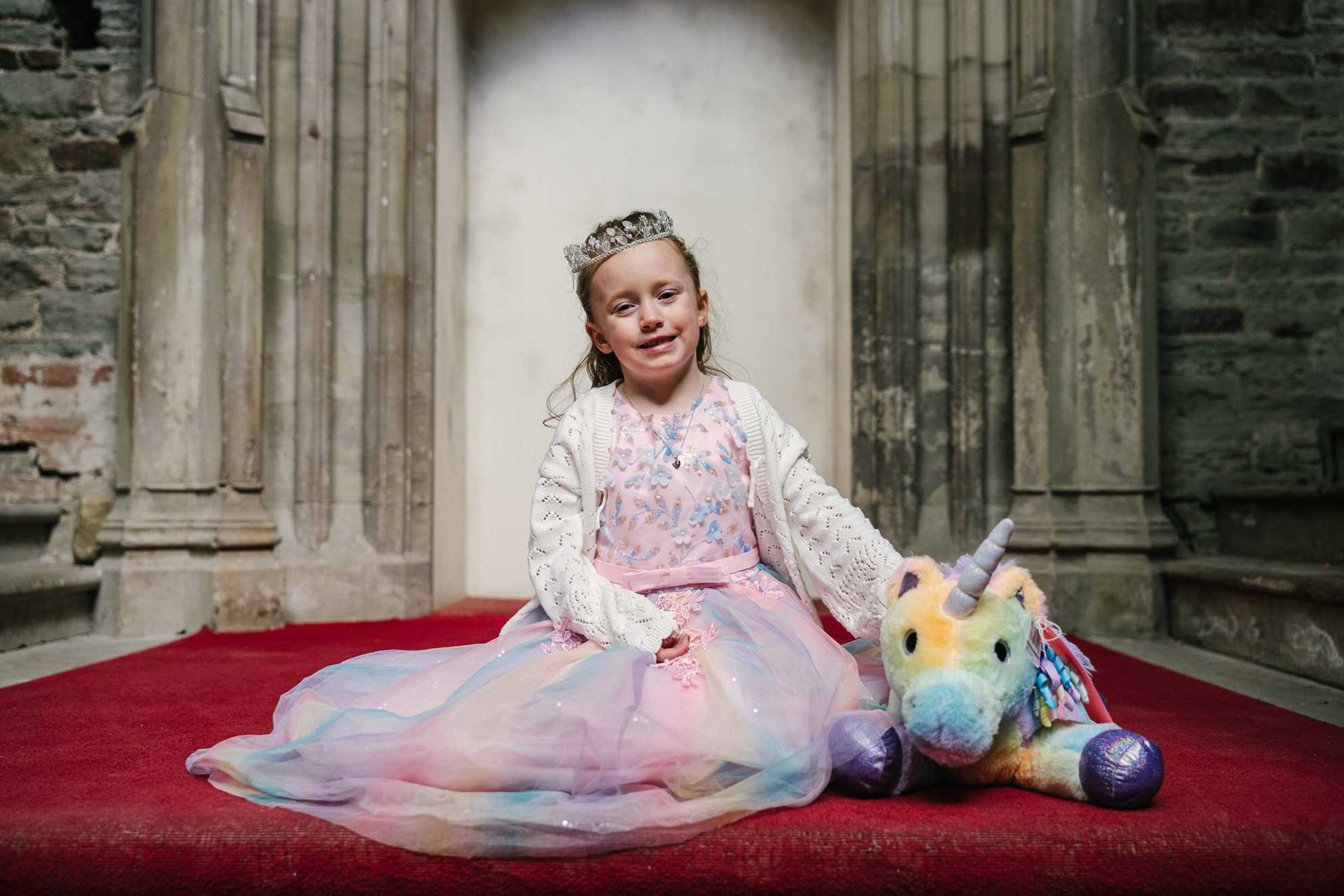 Penelope sits at the top of a staircase in a castle dressed as a princess with a toy unicorn alongside her
