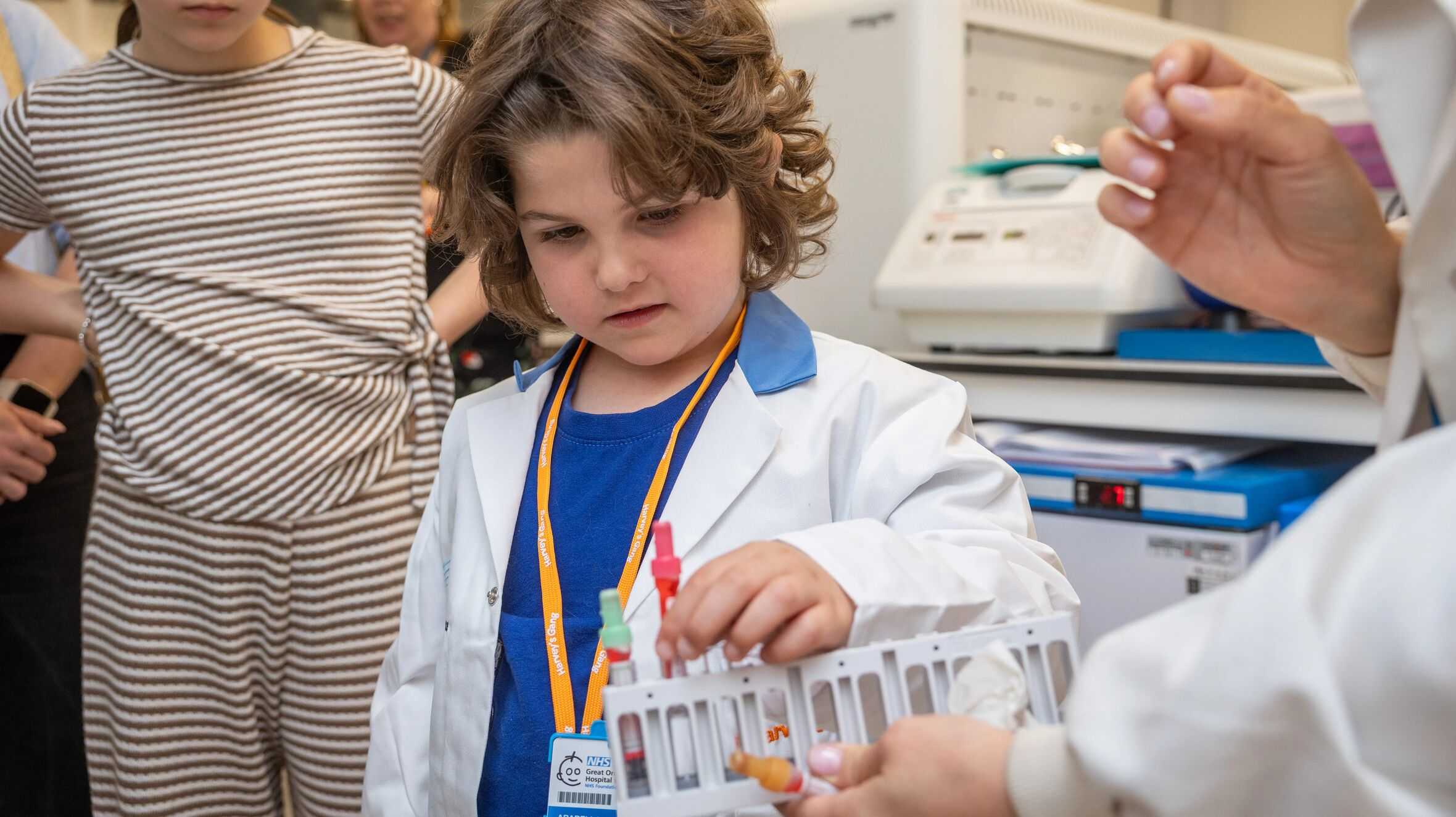 Arabella can be seen in a white doctor’s coat examining a vial rack. She is putting a small vial in the rack which is being held by an adult just out shot. Arabella is concentrating hard and appears to be taking her task very seriously.