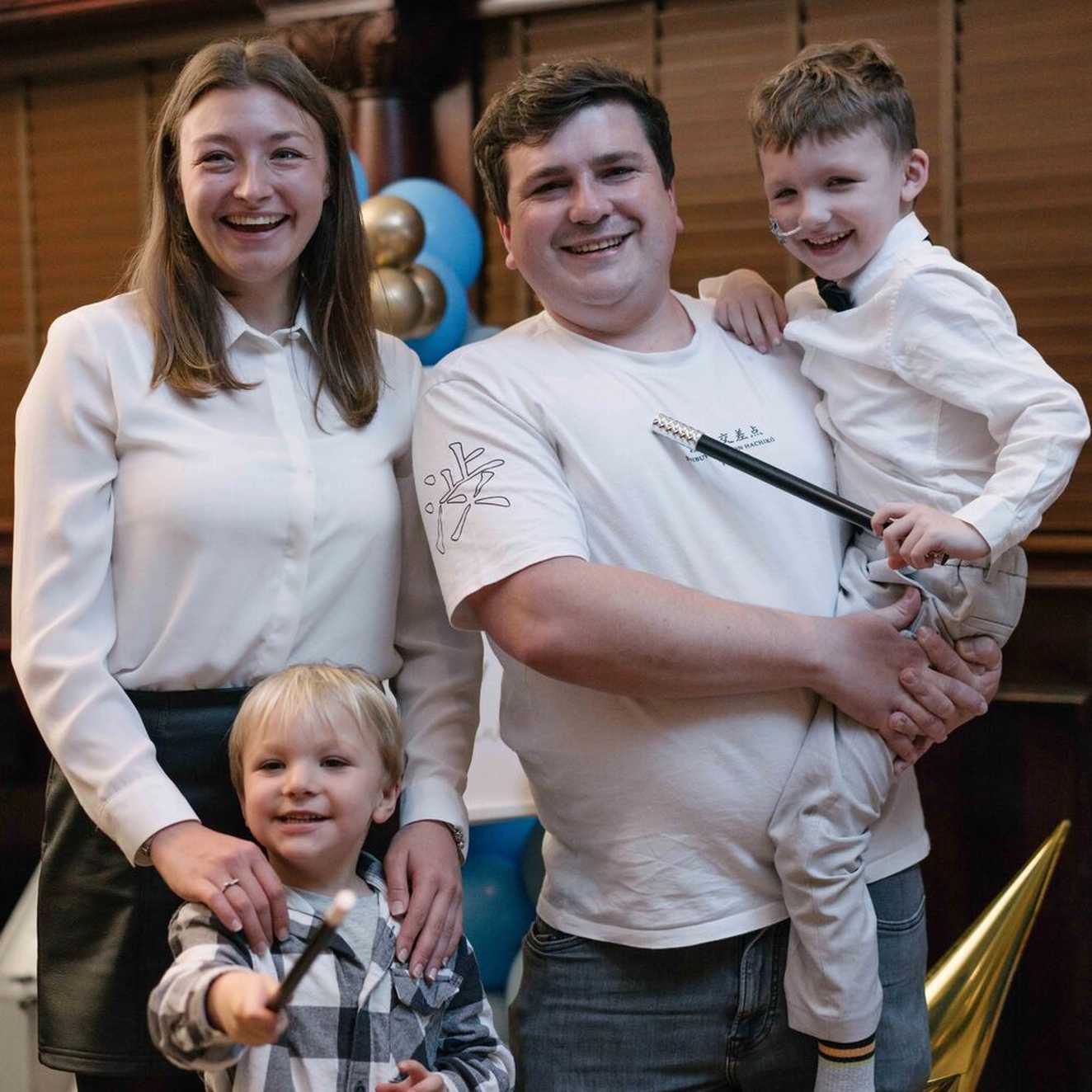 Charlie with his family, left to right, Mum, Brother and Dad.  His Dad is holding him and everyone is smiling.  Charlie and his Brother are holding magic wands.