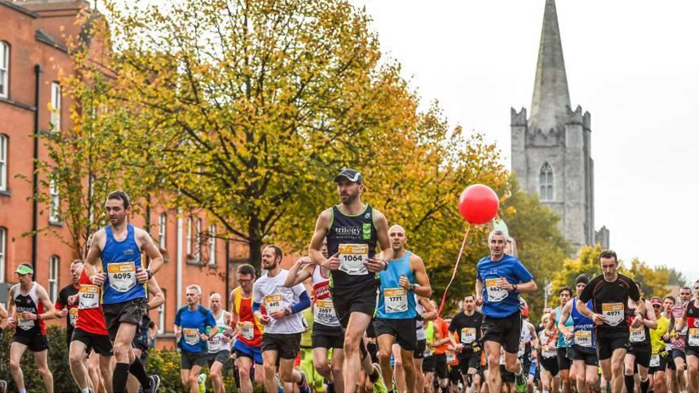 A group of Dublin Marathon runners, one carrying a red balloon, making their way through up a tree-lined street with a church steeple in the background.