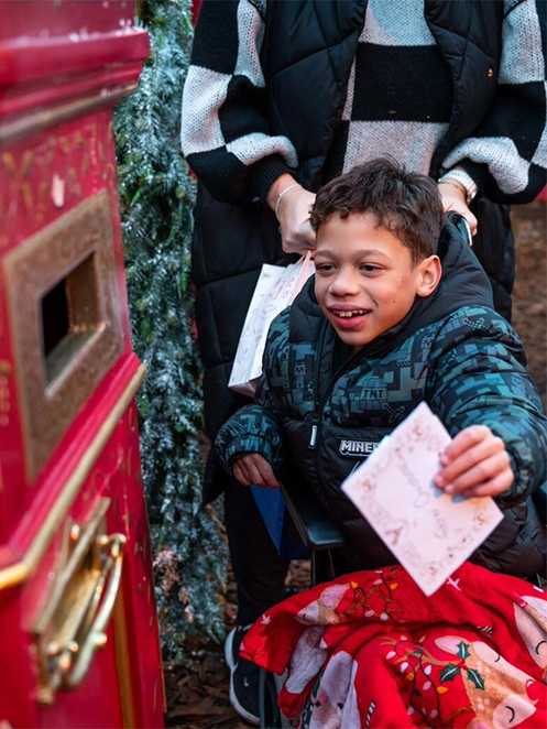 Kaiden is sitting in his wheelchair with a red Christmas blanket on his lap. He is reaching forward to post a letter to Father Christmas, in a red post box.