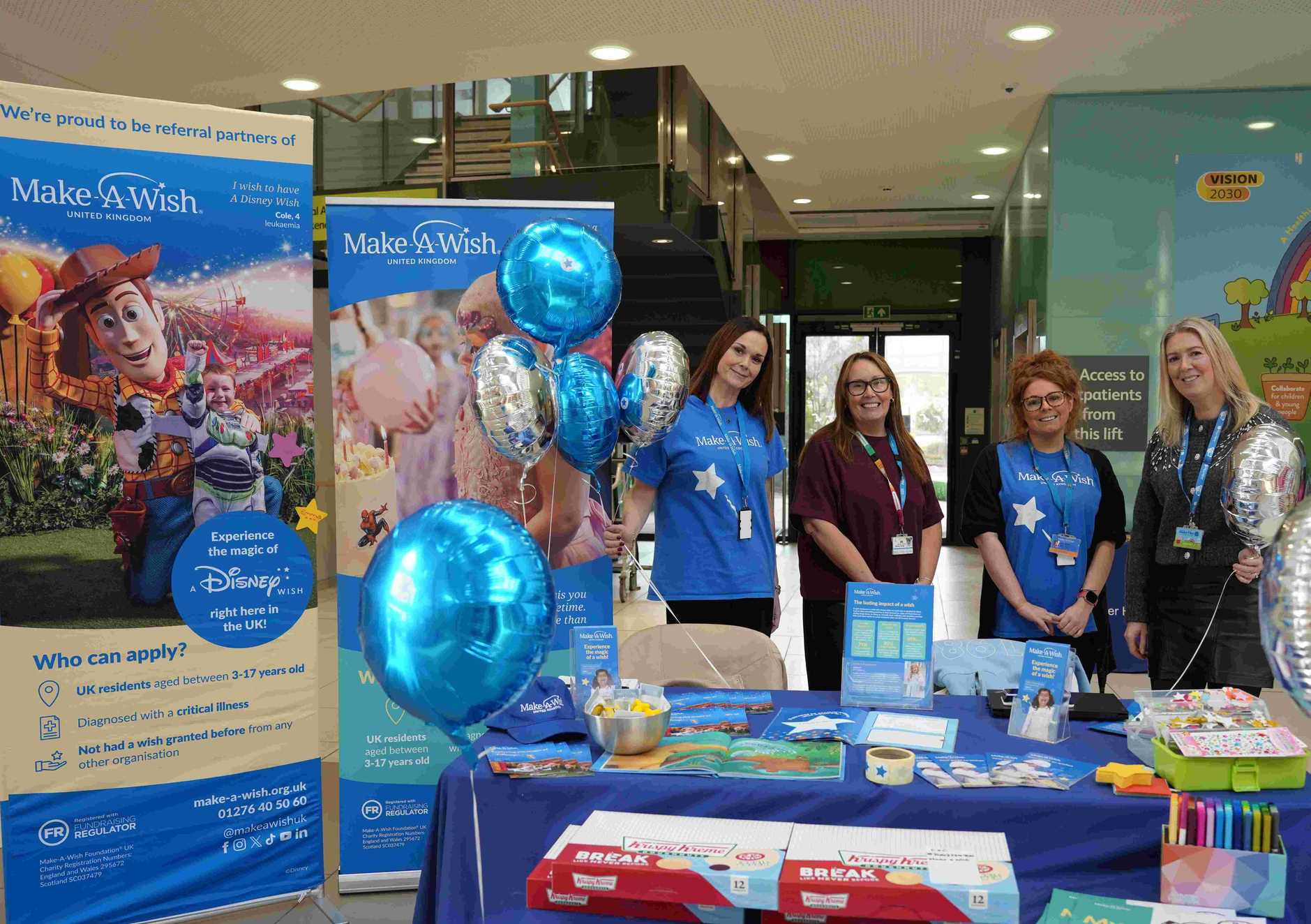 Indoor event display with four smiling women in Make-A-Wish shirts, surrounded by blue and silver balloons. Promotional materials cover a table. Bright, inviting atmosphere.