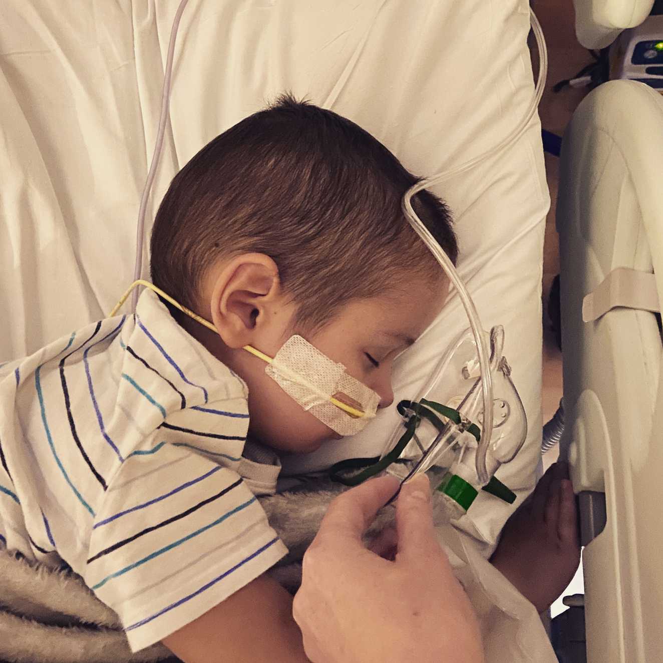 Liam lies asleep in a hospital bed during his treatment, with tubes and wires from his nose and around his head
