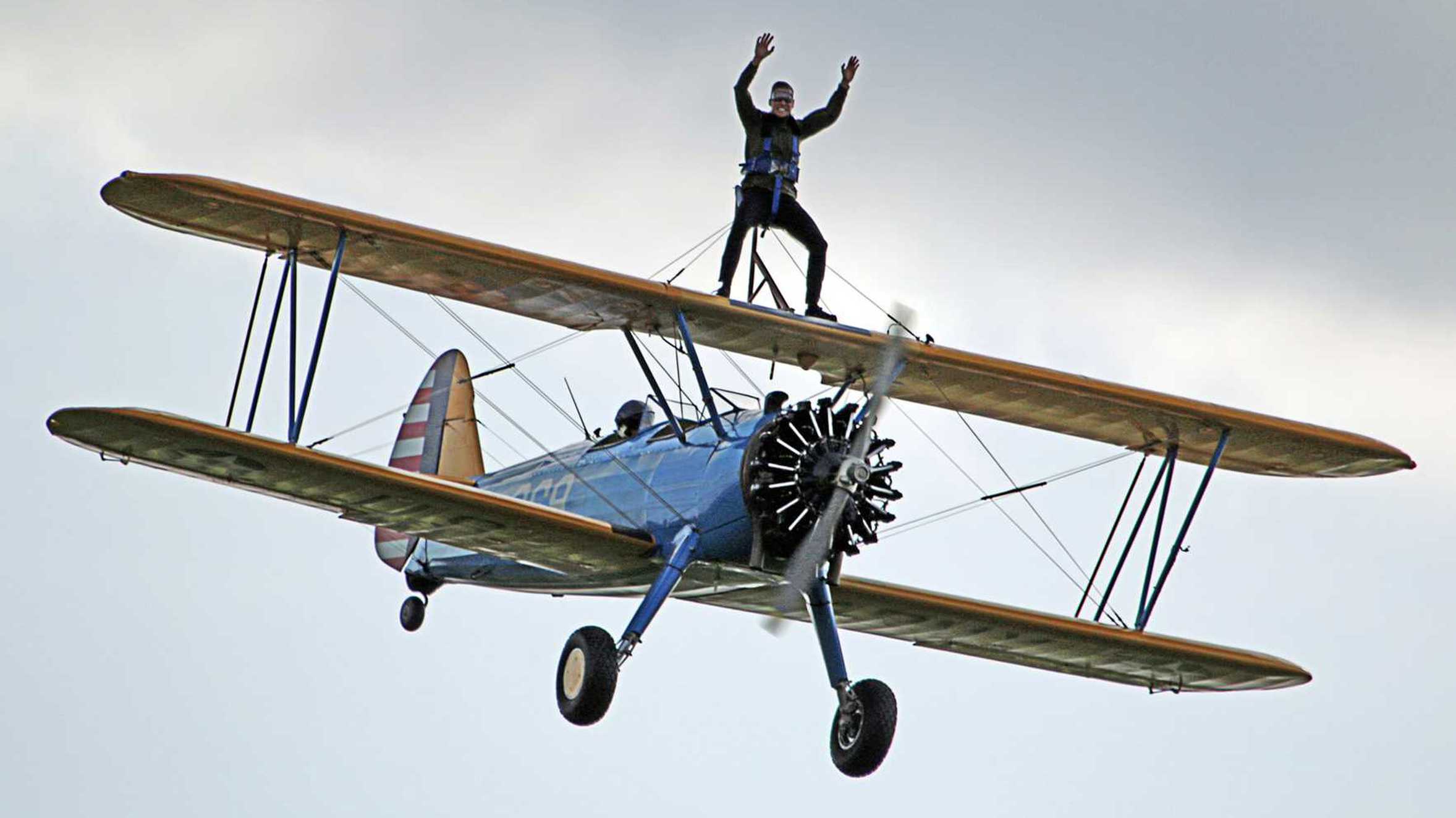 An intrepid supporter smiling while strapped to the wing of an airborne bi-plane.